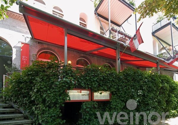 Rote Markise als Sonnenschutz vor einem Restaurant mit viel Grün und moderner Fassade.